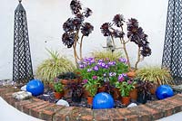 Raised corner bed contains pots of succulents, foliage and flowering plants, with decorative blue glazed ceramic spheres and metal obelisks - Bude Street, Appledore, Devon, UK