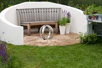 Wooden curved bench on circular patio with central sculpture and white rendered wall in the 'Ring the Changes' garden 