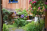 Wood and iron garden furniture next to modern wooden house. Plants include Clematis 'Vyvyan Pennell' and Rosa 'Rosarium Uetersen' 