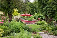 On one level of a big garden a rest area with wicker garden furniture, a parasol and a hammock is surrounded by flowering borders and old trees. Hedera helix, Matteucia struthiopteris, Phlox paniculata, Rosa and Salix integra 'Hakuro Nishiki'