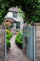 Open pale blue cottage garden gate with heart decoration, set into stone wall with Prunus laurocerasus 'Rotundifolia' archway cut in hedge, and pathway of stone sets leading to front door - Garden Neighbours