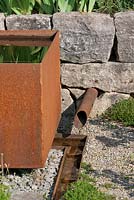 Next to corten steel water basin, a pipe coming out of a dry stone wall brings water to a rill