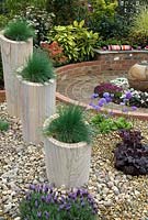 Trio of Festuca glauca 'Intense Blue' in tall stone planters on gravel border alongside circular patio, with contrasting shrubs in background - 'At the End of the Day' Show Garden, Silver Gilt Award, Harrogate Spring Flower Show 2013