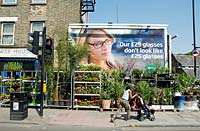 Urban garden centre in Mackenzie Road housed in the shell of an old building with mother and child passing the plants displayed on pavement and hoarding behind, Holloway, London Borough of Islington, UK