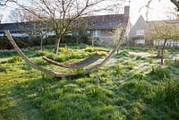 Orchard with wooden hammock set amidst meadow dotted with, camassias, Tulipa saxatilis, narcissi and snakeshead fritillaries, Fritillaria meleagris. Forest Lodge, Pen Selwood, Somerset, UK