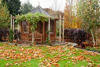 Autumnal garden with wooden Summer house. Planting of Fagus sylvatica 'Dawyck Gold', Betula utilis jacquemontii 'Doorenbos', Trachelospermum jasminoides and Wisteria sinensis 'Prolific'