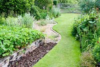 Curving lawn leading to gate by herbaceous border, raised vegetable bed with strawberry plants and graveled area in cottage garden