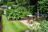 Graveled seating area with bench and raised vegetable beds in cottage garden surrounded by mature trees