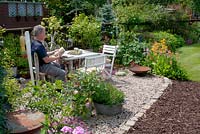 Man seating in graveled corner of cottage garden with small table and chairs and collection of vintage galvanised containers - planting includes; Rosa, Primula, Argyranthemum and Geranium 