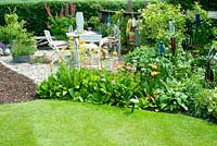 Seating area with small square table and chairs made of drift wood on graveled corner of cottage garden with collection of vintage galvanised containers fishing floats and planting including Lupinus, Primula and Geranium 