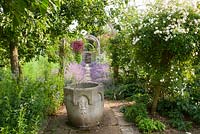 A gravel and stone set path edged with Nepeta 'Six Hills Giant' runs through the centre of the potager garden with timber archway marking its centre supporting clematis, roses and Trachelospermum jasminoides, with stone font in foreground. 