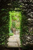 Collection of containers of red flowering plants framed by opening in yew tunnel. 