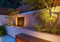Minimalist garden lit up at night with built in raised beds - Edgeworthia Chrysantha, Acer Aconitifolium and water feature 