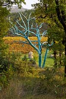 View through the woodland to vines and a stone well in front of a tree painted blue by Marc Nucera. Provence, France, Domaine de la Verriere
