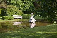 Hokusai's Boat sculpture in tranquil pond garden with clipped hedging and wooden seat.  Farleigh House, Hampshire