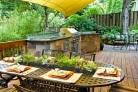 Summer outdoor kitchen with grill and sink on wooden deck. Wrought-iron chairs and table with succulent garland centerpiece.