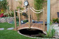 Ornamental wooden bridge over small pond to patio, clock, metal spheres and brick raised bed in the 'Thyme on our Hands' garden by B G Fencing Ltd - Southport Flower Show 2013