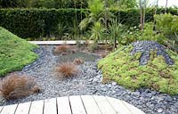 Title: Pour L Amour de Tongariro. Garden with wooden path passing several 'volcanos' planted with sedums and covered with broken black slates.
