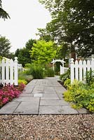 Flagstone garden path leading to arbour through a white picket fence. Planting includes purple caucasian stonecrops (Sedum spurium), yellow Lady's mantle (Alchemilla mollis), Kleine 'Silberspinne' (Miscanthus sinensis) in the middle and a black locust (Robinia pseudoacacia 'Frisia') tree in the background. Il Etait Une Fois garden, Monteregie, Quebec, Canada. 