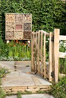 Feature the Visible garden. Rustic split cane gate opens on to stone flooring with wooden insect hotel set against hornbeam hedge.  Wildlife garden   