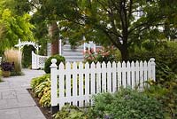 Grey flagstone path leading to a arbour through a white picket fence in front garden in autumn. Plantings include Alchemilla mollis and a Maackia amurensis tree. Stacked log home in the background. Il Etait Une Fois garden, Monteregie, Quebec, Canada. 