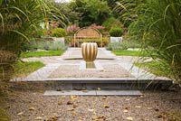 Raised gravel bed with gourd-shaped water fountain and brown metal lattice garden bench with pink hydrangea paniculata 'Quick Fire' framed by tall Miscanthus sinensis 'Berlin' ornamental grasses in backyard garden in autumn. Il Etait Une Fois garden, Monteregie, Quebec, Canada. 