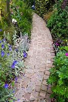 Curved cobble setts path softened by planting of Geranium 'Orion', Stachys byzantina, Coronaria and Heuchera 'Obsidian' with mirrored water feature nestled in amongst the planting