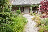 A path through the front garden is lined with grasses, euphorbias, bamboos, ferns, clipped box and a dark red acer. Plain paving is broken up by a lozenge shape of inset pebbles. 