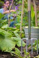 Garden fork and metal bucket in a raised bed, with woman working in background