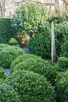 A topiary garden filled with low clipped box bushes surrounded by gravel, a slender standing stone and standard Portuguese laurels. 