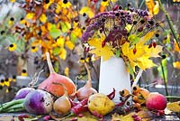Jug of perennials and autumn leaves and harvested vegetables - rosehips, Verbena bonariensis, maple, Rudbeckia triloba, Sedum 'Herbstfreude', onions, apples, pears, squashes, turnips, peppers.