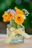 A summer posie of everyday garden flowers - Nasturtium, Aster and Anthemis in a glass jar decorated with yellow gingham ribbon.