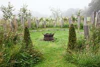 Wild meadow planted around a circular firepit on a misty morning, with oak obelisks and Buxus pyramids.