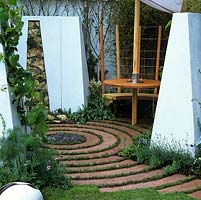 Tiny, modern circular courtyard with central slate mound. Alternating circles of grass and concrete. White, slab walls contain dry stone panels. Marine style dining area.