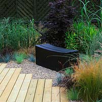 Simple block of black, curving wood creates seat in gravel garden amidst grasses, bamboo and red acer.