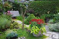 Pink Astilbes next to a footbridge and grey painted wooden swing bench underneath a pergola decorated with red Fuchsia flowers in a hanging basket and red Pelargonium - Geraniums with Ipomoea batatas in planter in backyard garden in summer, Jardin Secret garden, Quebec, Canada