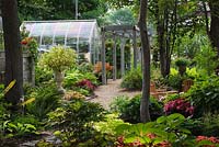 Cedar mulch path leading to greenhouse and gray painted wooden pergola underplanted with orange Begonia boliviensis 'Santa Cruz' flowers and Cimicifuga racemosa - Cohosh and Rodgersia plants in foreground in backyard garden in summer, Jardin Secret garden, Quebec, Canada