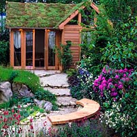 Sloping garden with stepped gravel path. Shed with tumbling green sedum roof. Curved wood bench by rhododendron, seen through cirsium, verbascum, valerian and aquilegia.