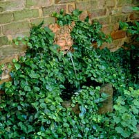 A mythical face, cast in clay and set in ivy, spouts water into a small trough.