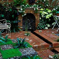 Tudor bricks laid in a herringbone pattern frame a rill leading to a flint alcove where water falls, set under a mantel. Floor made from alternating squares of chamomile, agave and flint.
