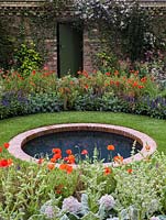 Circular pool in round lawn edged in beds of poppies, eryngium and salvia. Beyond, wall clad in Clematis montana 'Rubens' with door.