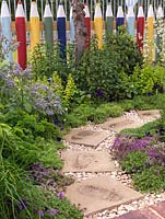 Fun picket fence in shape of coloured pencils edges herb bed of mint, borage, parsley, thyme, chives, oregano, chamomile and strawberry. Paving stones through gravel.