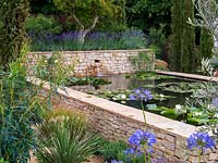 Raised pool, clad in dry stone walls, is home to water lilies. Mediterranean style garden with lavender, agapanthus, cypress, olive, oleander, rosemary and restio.