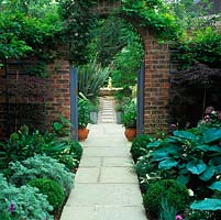 At side of house, paved path - edged in hosta, box, santolina and Allium cristophii - leads to arch framing view of path continuing to fountain. Creates illusion of greater space.