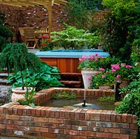 Outdoor hot tub is edged in geranium, hosta and photinia, partly concealed behind raised brick pool. Above, outdoor seating area beneath pergola.