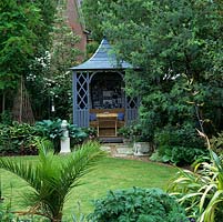 Seen through palm, phormium and hardy geranium, circular lawn with summerhouse enclosed in acacia, contorted willow, maple and robinia for privacy. On left, shady hosta bed.