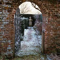 In ancient brick wall, iron gate opens to reveal frosty path leading to bust of Prince of Denmark set against yew hedges.