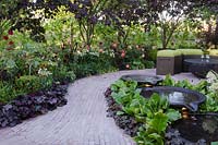 Circular pools next to a brick path, leading to a seating area with green cushions, dark contrasting foliage of Cercis canadensis 'Forest Pansy' and Heuchera 'Black Beauty', 'Plum Pudding' and 'Chocolate Ruffles' - Bacchus Garden, RHS Hampton Court Palace Flower Show 2014 