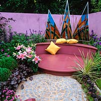 Triangular triptych of stained glass rises above interlocking patios of circular granite - garnet coloured decking. Pink wall echoes lilies, heuchera, salvia, hardy geranium and pinks.