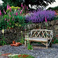 Sunken stone garden with wooden bench, white foxglove and red Geum 'Mrs Bradshaw' self-seeded in slate. On wall, Nepeta 'Six Hills Giant' and foxgloves.